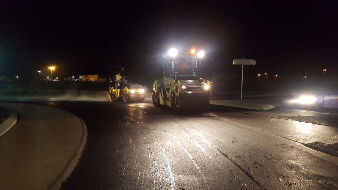 Travaux de pavage nocturnes ; un rouleau compresseur et un ouvrier compactent l’asphalte sur une chaussée.