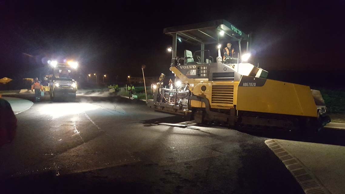 Travaux routiers de nuit. Finisseur et rouleau compresseur compactant l'asphalte. Ambiance sombre, éclairée par des projecteurs de chantier.