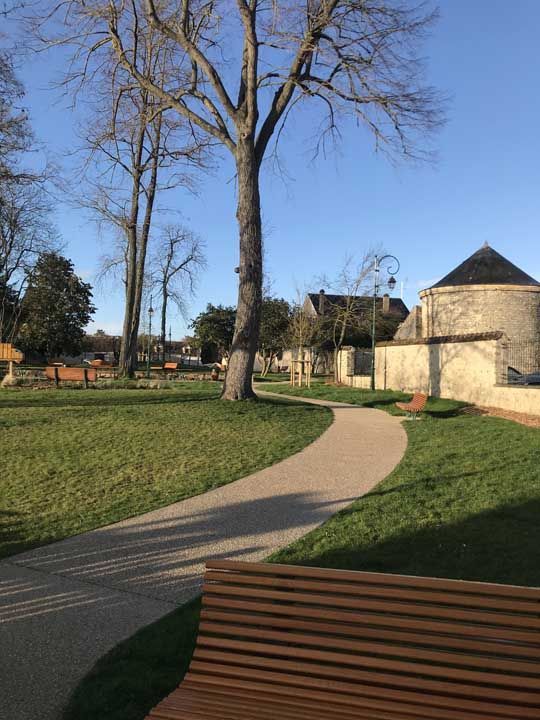 Parc avec un chemin sinueux, des espaces verts, des bancs en bois et des arbres dénudés sous un ciel bleu limpide.