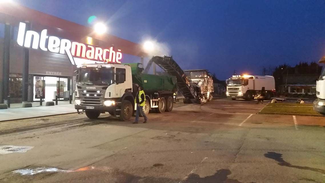 Des engins de chantier effectuent le pavage d'un parking devant un supermarché Intermarché au crépuscule.
