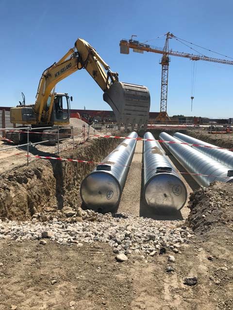 Une excavatrice place des réservoirs cylindriques dans une tranchée sur un chantier de construction ; une grue est visible en arrière-plan.