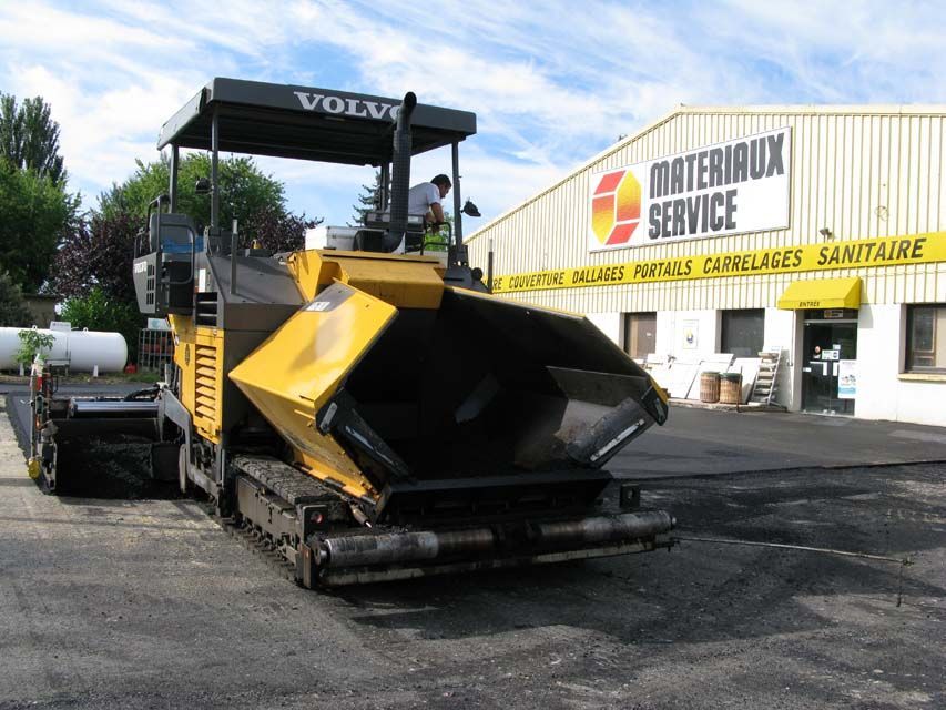 Une machine à asphalter Volvo jaune pose du nouvel asphalte devant un bâtiment.