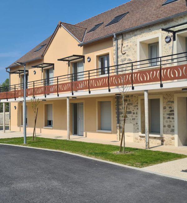 Bâtiment de deux étages aux murs beiges, orné de détails en pierre et d'un toit brun. Un balcon au deuxième étage est doté de balustrades décoratives couleur rouille.
