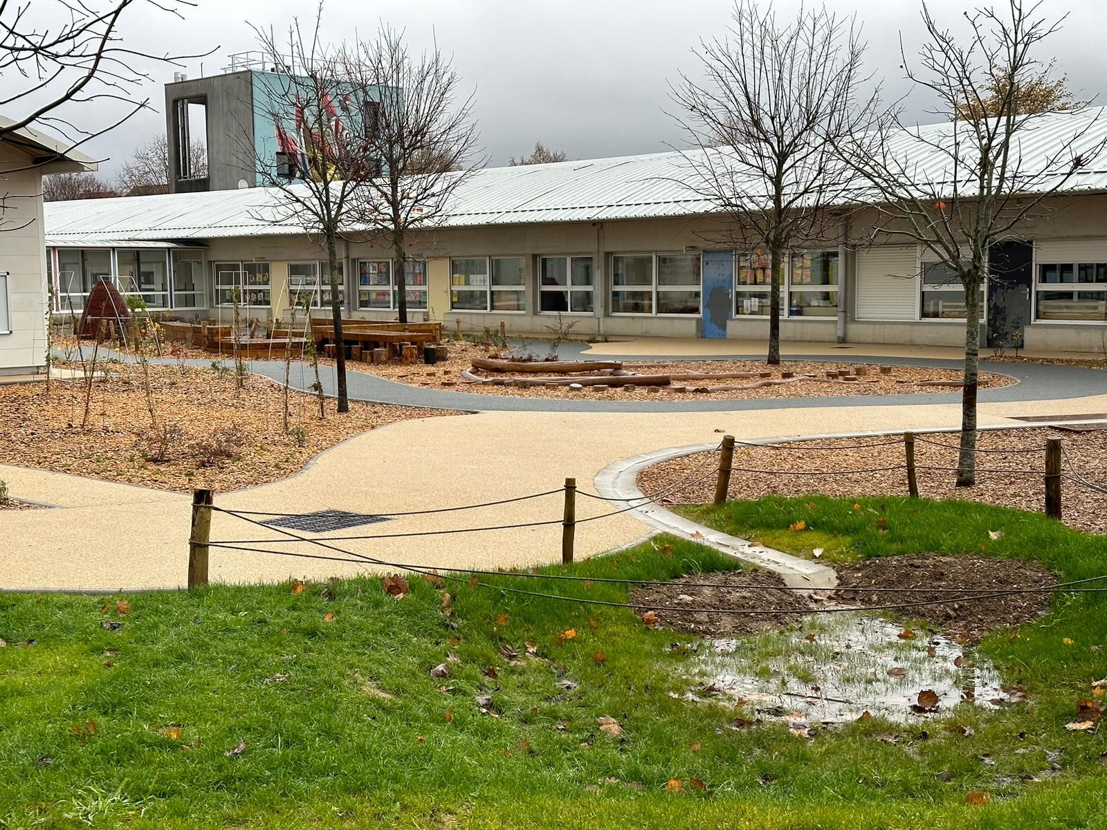 Bâtiment scolaire avec un chemin de gravier, un petit étang et des arbres au premier plan. Ciel couvert.