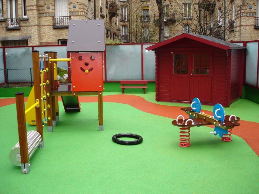 Aire de jeux avec des structures rouges et marron sur un sol vert et orange, un pneu noir et un abri rouge.