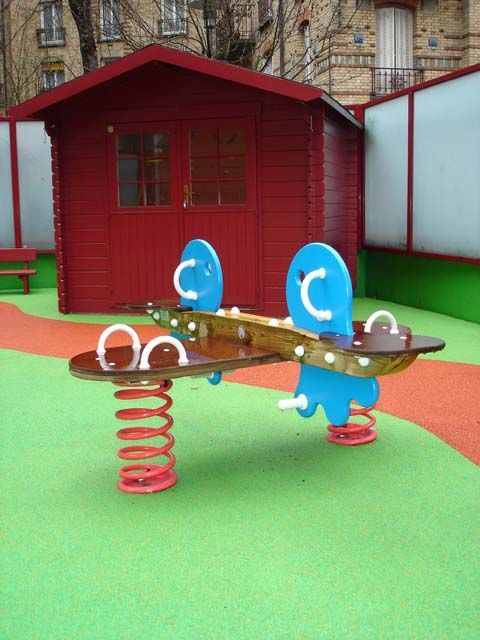 Maisonnette au toit rouge et balançoire bleue à ressort sur un sol de jeu vert et orange.