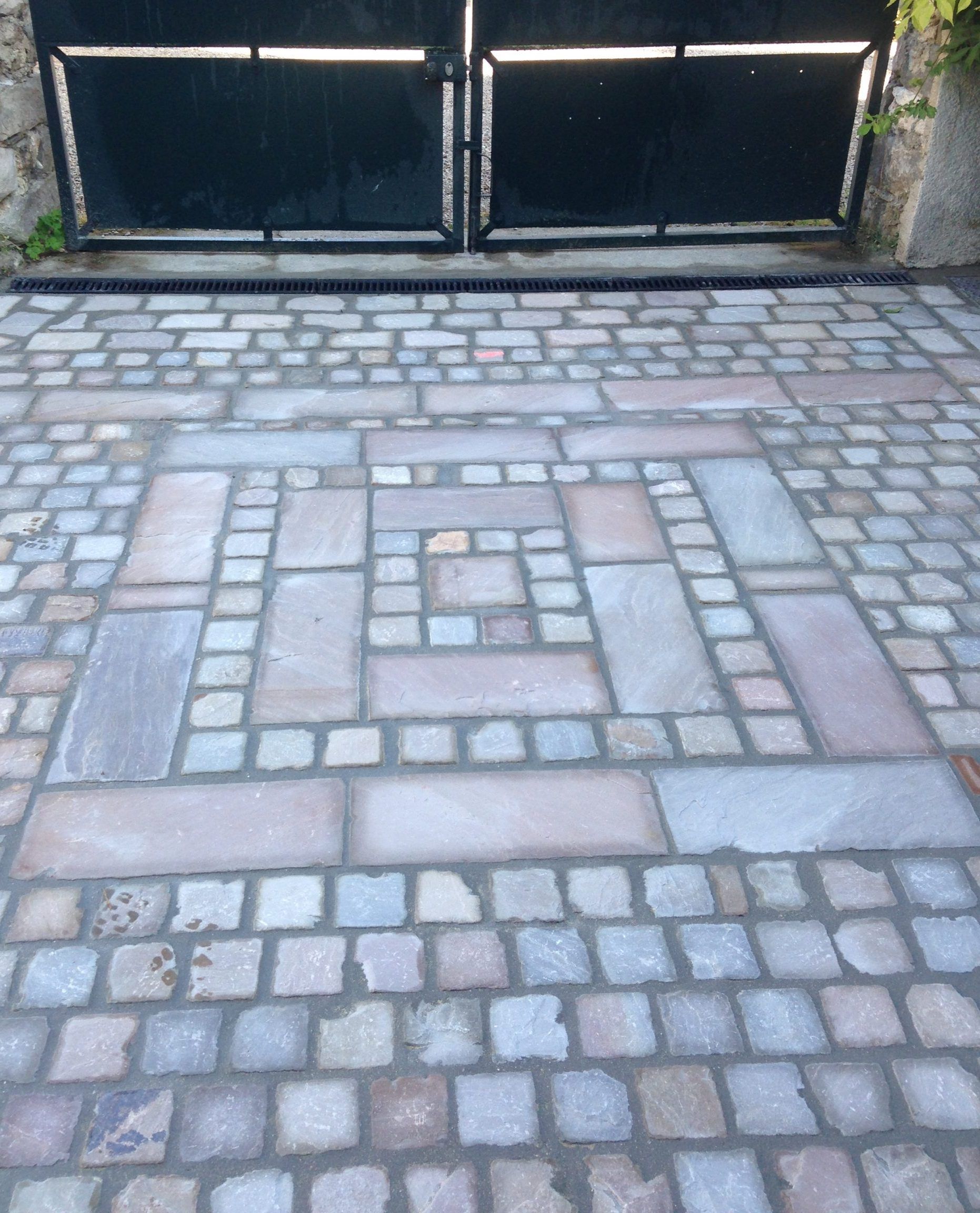 Allée pavée à motif géométrique menant à un portail en métal noir.