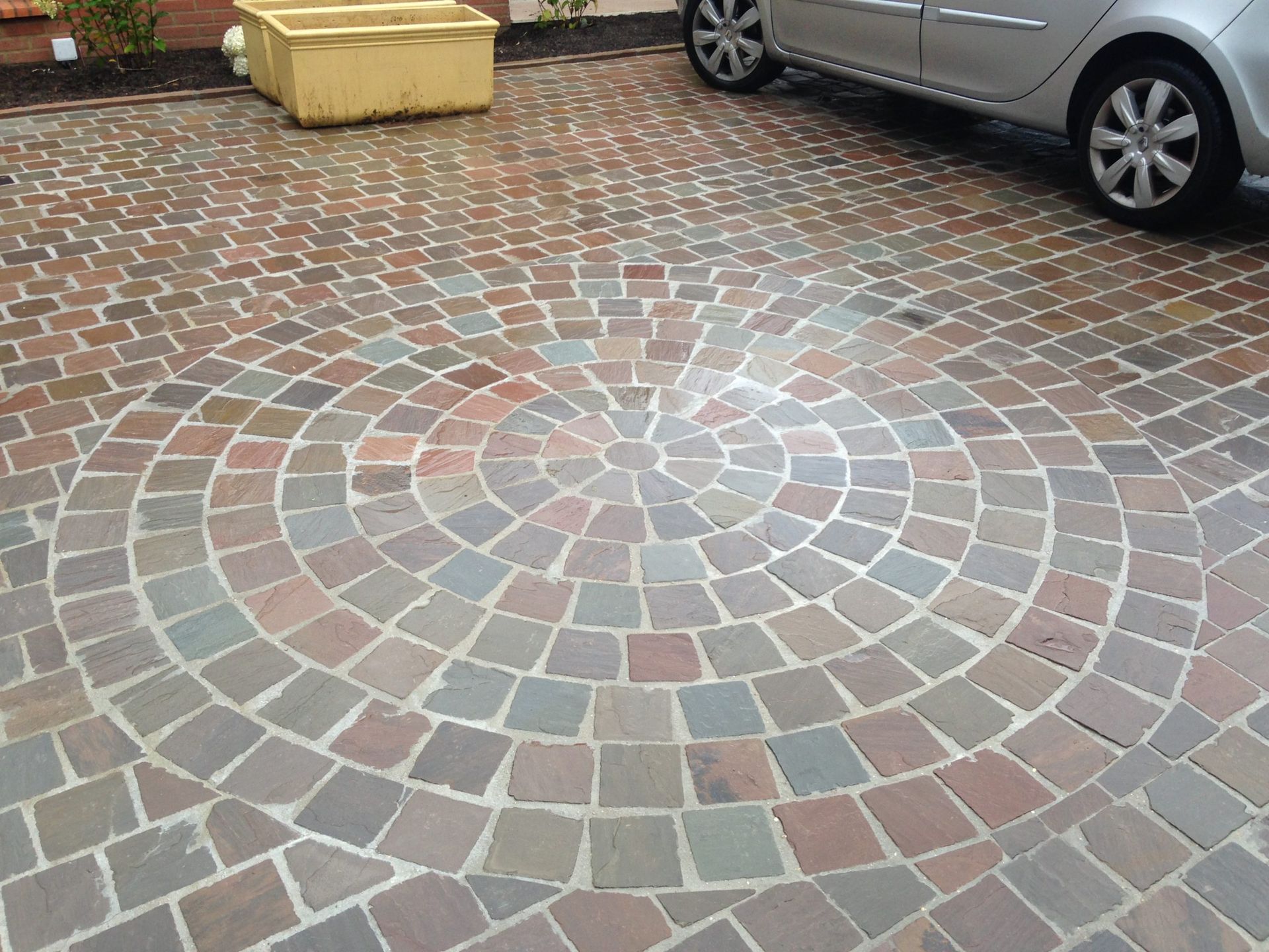 Allée pavée de briques circulaires ; voiture garée, poubelle jaune.