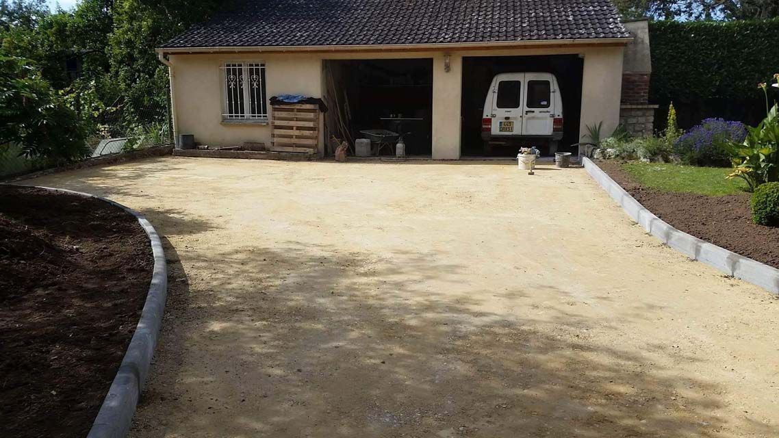 Allée en gravier beige menant à un garage avec une camionnette blanche. Bordures paysagées avec terre foncée et bordures grises.