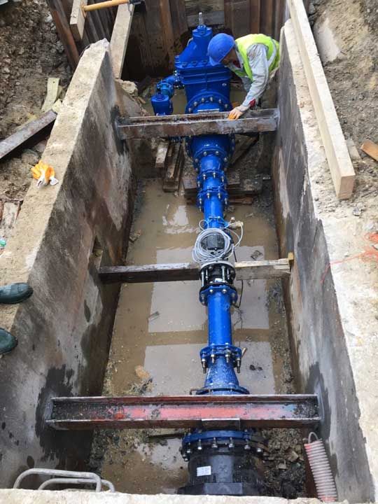 Un homme portant un casque de chantier travaille sur une grosse conduite d'eau bleue dans une tranchée en béton.