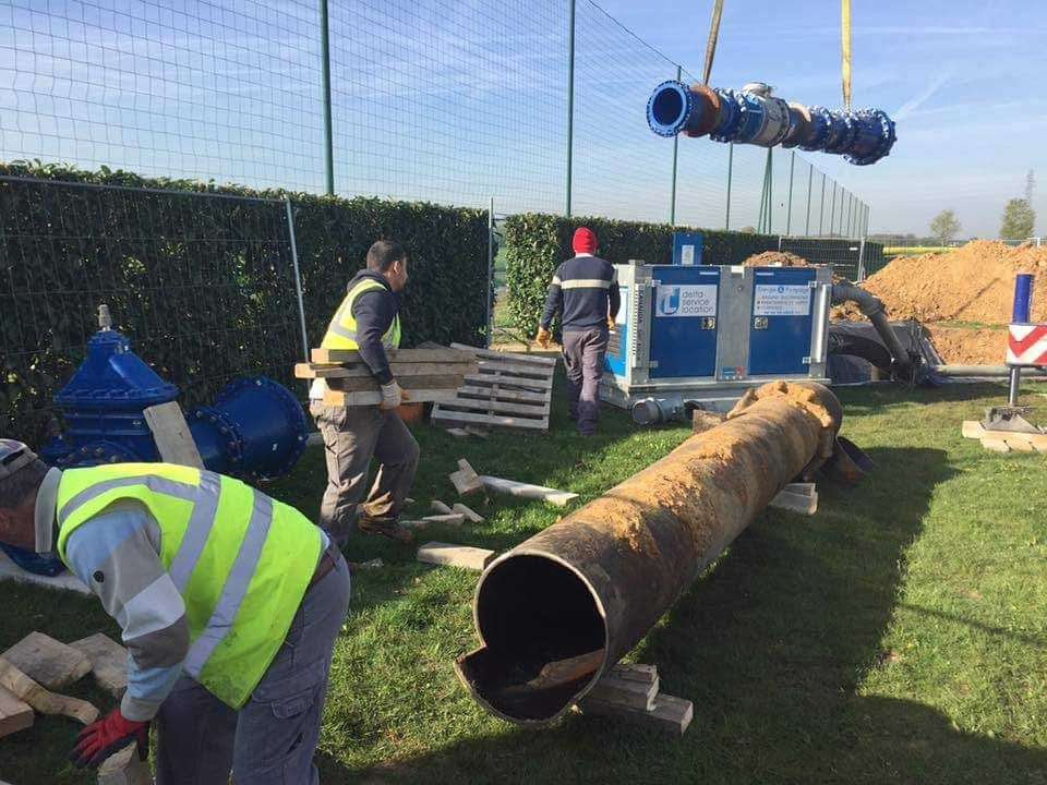 Chantier : ouvriers installant des canalisations. Une grosse canalisation au sol, une autre suspendue par une grue. Hommes en gilets. Clôture verte, ciel.