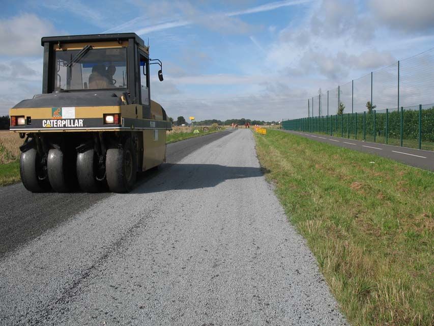 Rouleau compresseur compactant une route de gravier ; champ et clôture en arrière-plan.