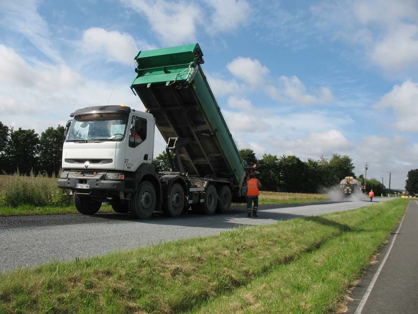 Camion-benne déchargeant du gravier sur une route ; ouvriers du bâtiment portant des gilets de sécurité ; journée ensoleillée.