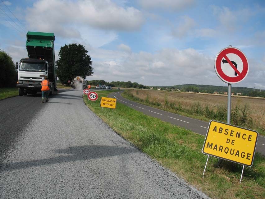 Réfection de la chaussée : camion déversant de l’asphalte sur une route, ouvrier à proximité, panneaux de signalisation sur un bas-côté herbeux, champs au loin.