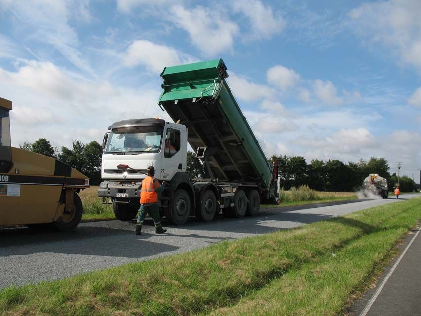 Un camion déverse de l'asphalte sur une route en cours de pavage ; un ouvrier en gilet orange donne les instructions.