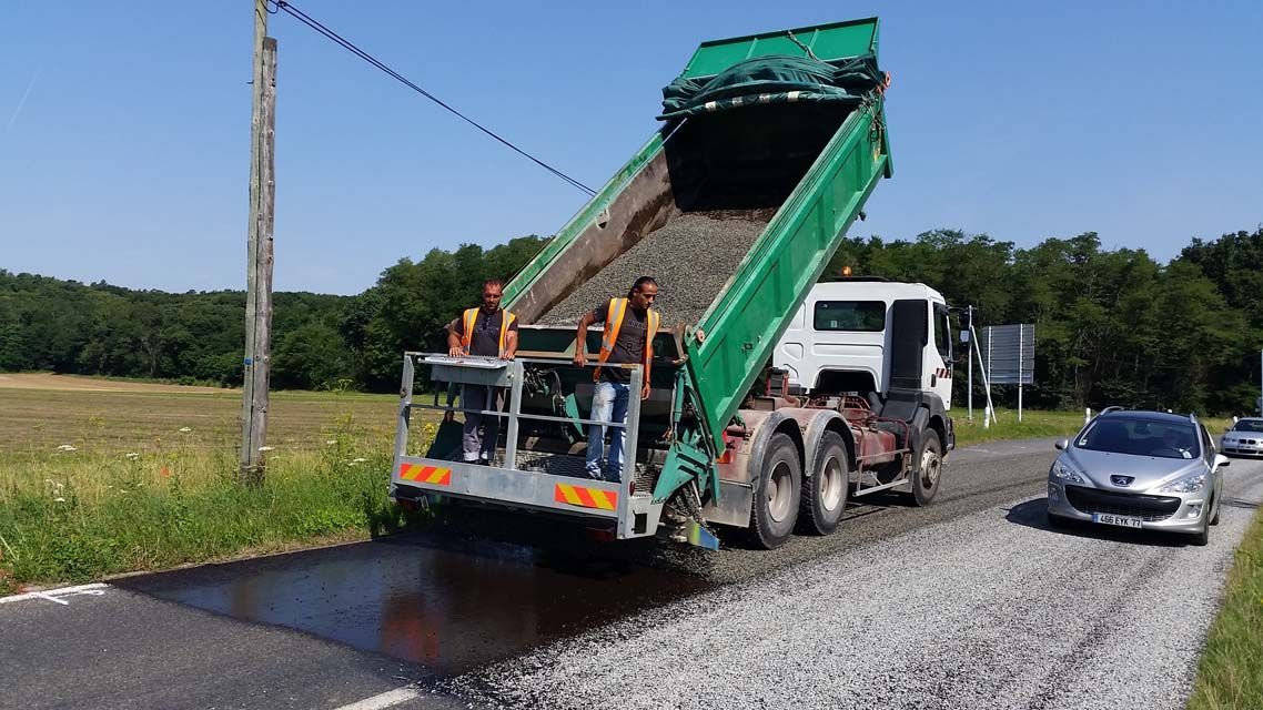Travaux routiers : un camion déverse du gravier sur une route asphaltée, deux ouvriers observent. Ciel bleu, voitures à proximité.