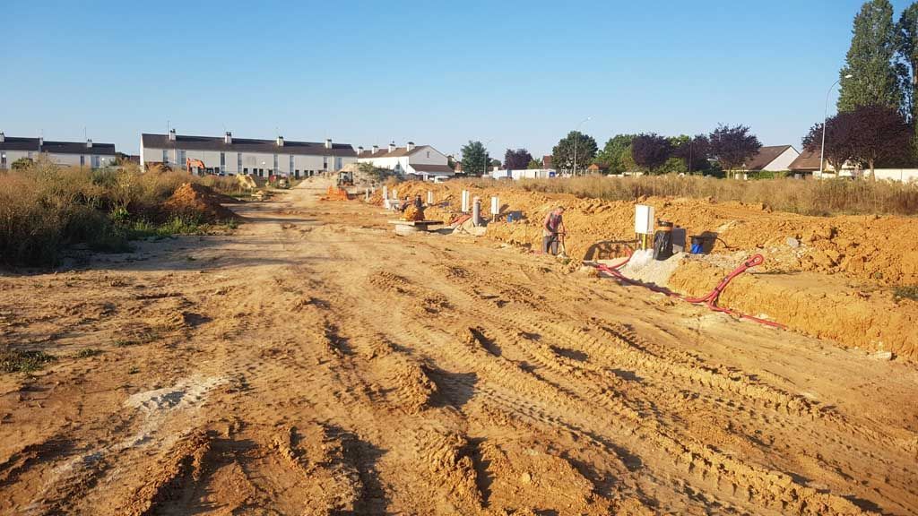 Chantier avec chemin de terre et fondations en béton. Maisons en arrière-plan sous un ciel bleu.