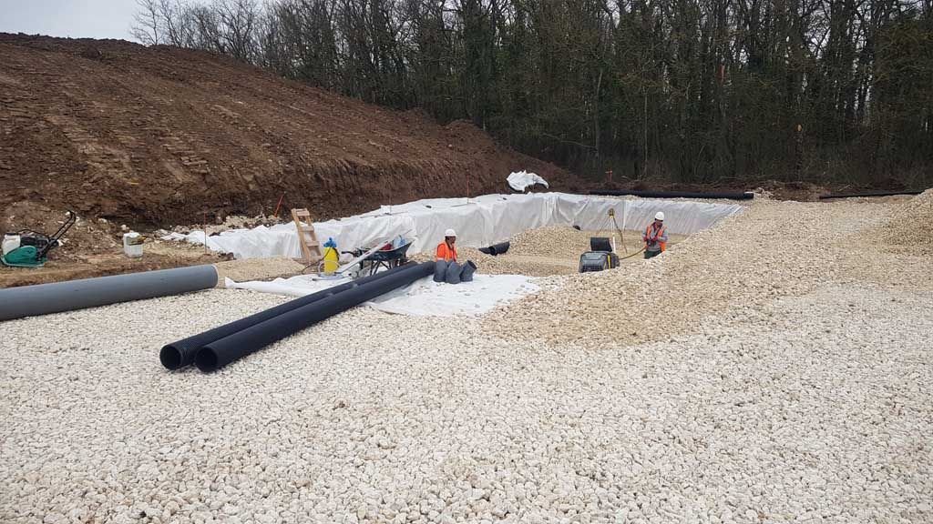 Chantier de construction avec des tuyaux, du gravier et des ouvriers près d'une butte de terre et d'arbres.