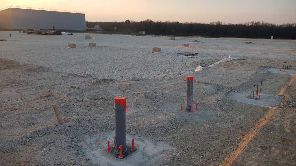 Chantier de construction avec sol en gravier et piliers en béton à capuchons orange.