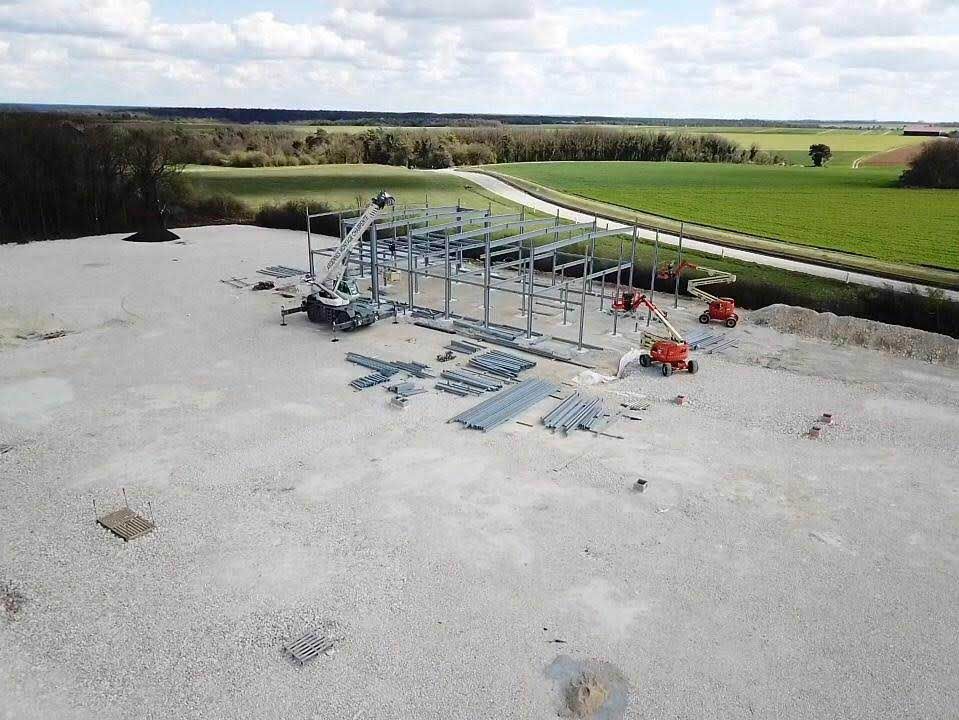 Chantier de construction : bâtiment à ossature métallique, machines, terrain gravillonné, champ en arrière-plan.