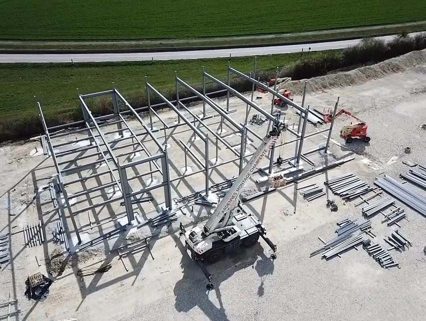 Chantier de construction avec charpente métallique en cours d'érection ; grue et engins de chantier présents.