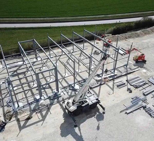 Charpente métallique d'un bâtiment en construction, avec une grue sur une dalle de béton et une nacelle élévatrice sur la droite.