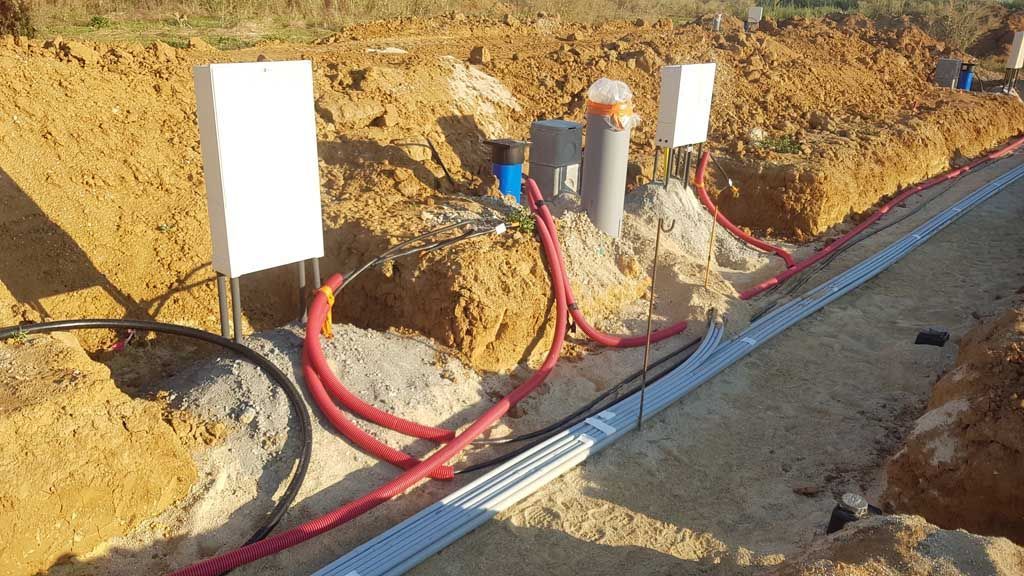 Tranchée avec conduits et tuyaux électriques, y compris des câbles rouges et noirs, à proximité de boîtiers blancs, dans une zone excavée.