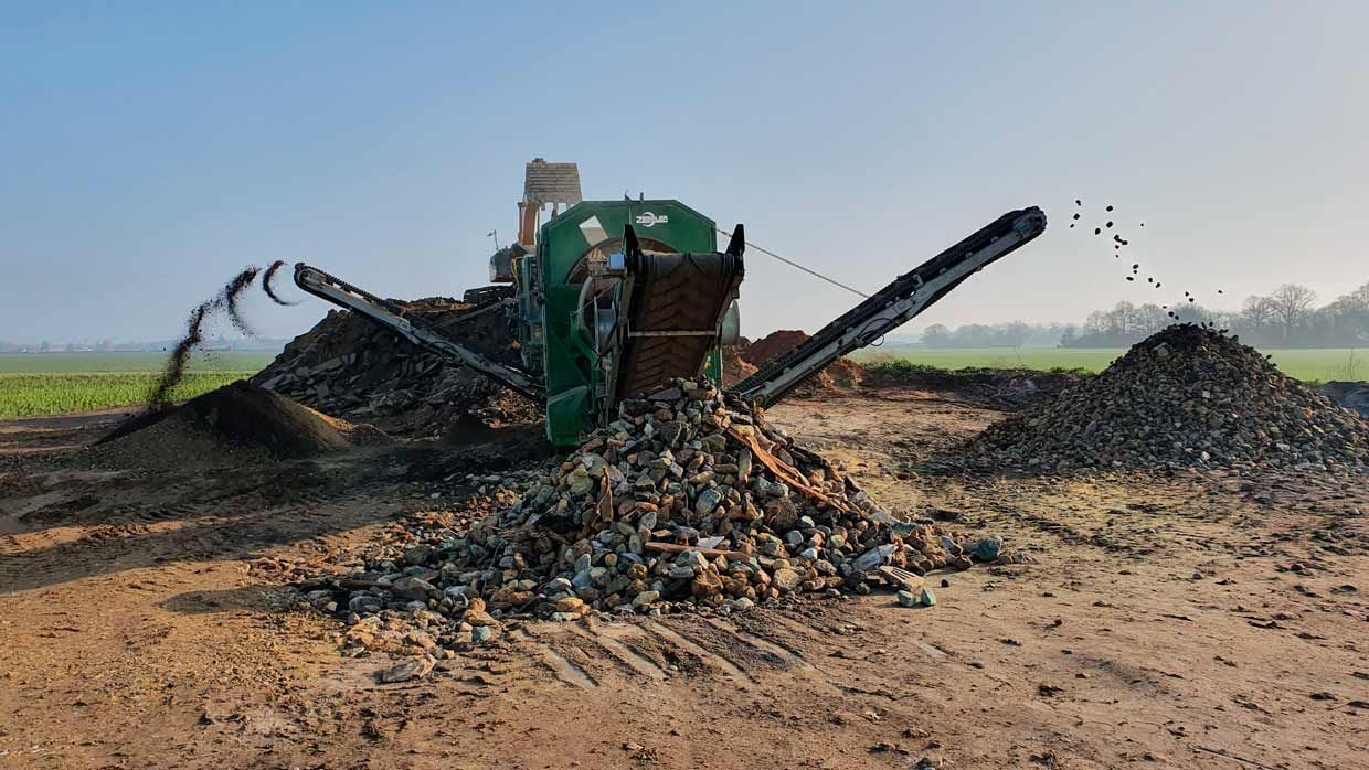 Une machine à concasser les pierres, crachant des roches sur un tas dans un champ. Ciel bleu en arrière-plan.