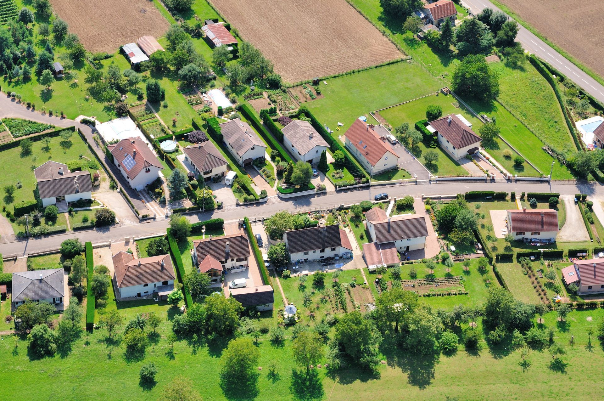 Vue aérienne d'une rue rurale tranquille bordée de maisons individuelles entourées de pelouses verdoyantes, de jardins et de terres agricoles.