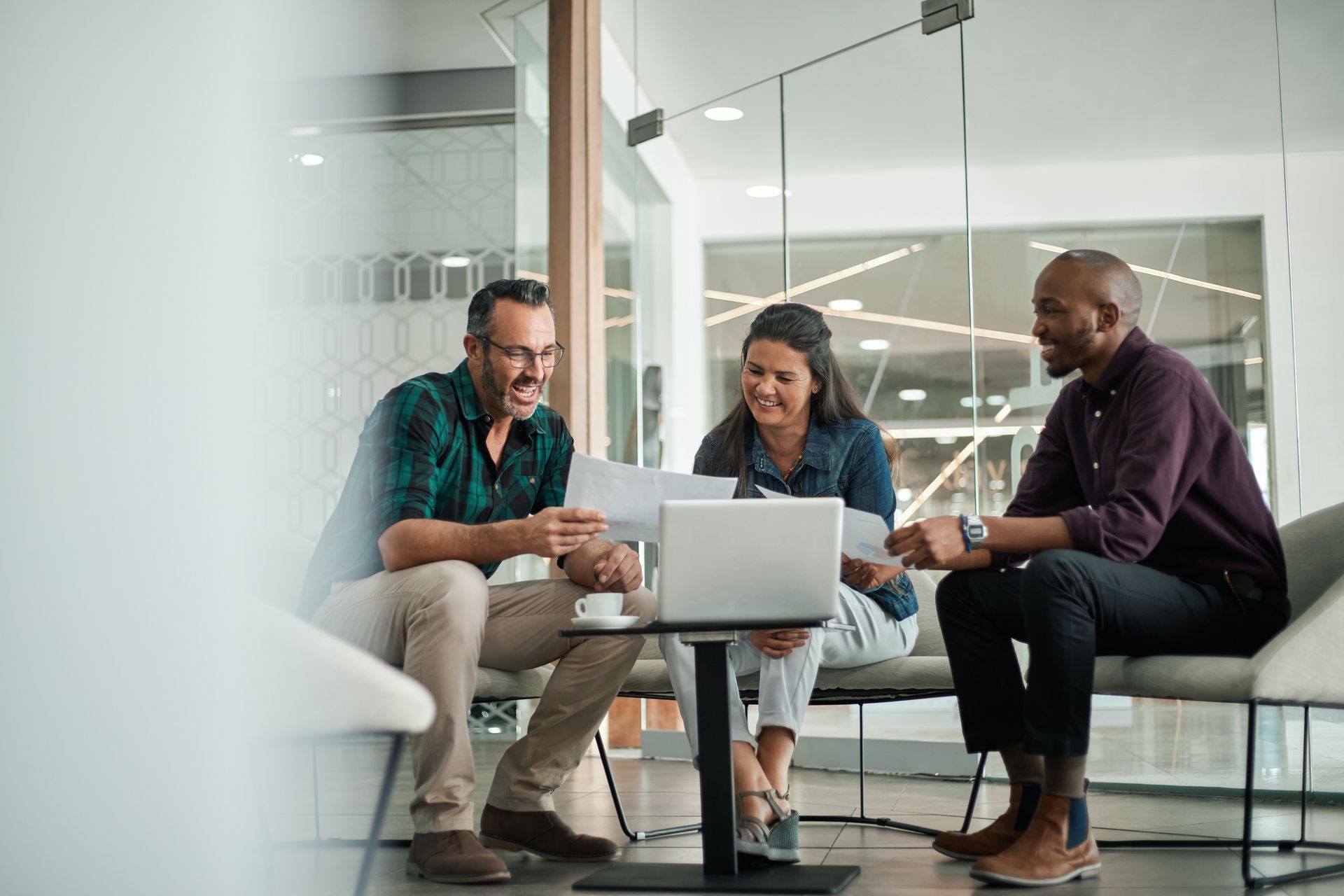 Trois collègues sont assis ensemble dans un bureau lumineux, souriant tout en discutant de documents et d'un ordinateur portable.