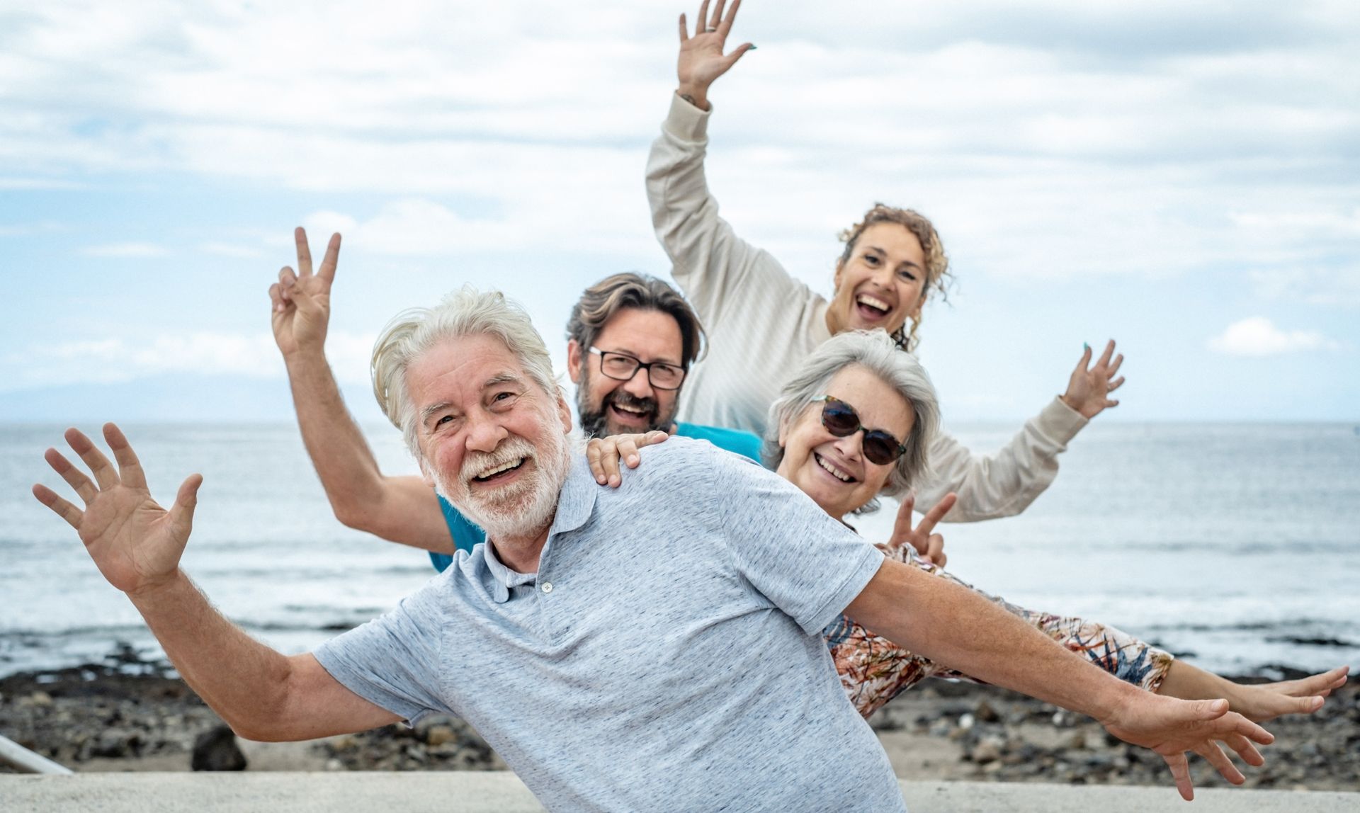 Quatre personnes posent de manière enjouée sur la plage, les bras tendus, souriant devant un ciel nuageux et l'océan en arrière-plan.
