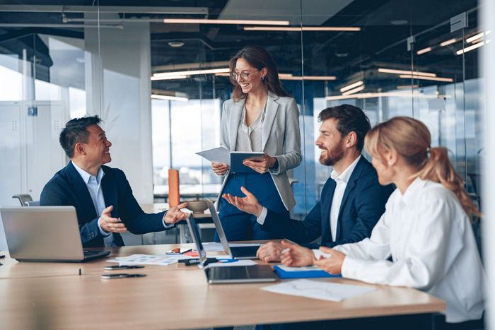 Quatre professionnels réunis dans un bureau moderne, discutant de travail assis et debout autour d'une table avec des ordinateurs portables.