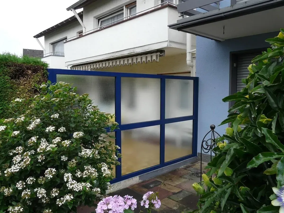 Blau gerahmter Zaun aus Milchglas vor einem Haus, neben einem blühenden Busch.