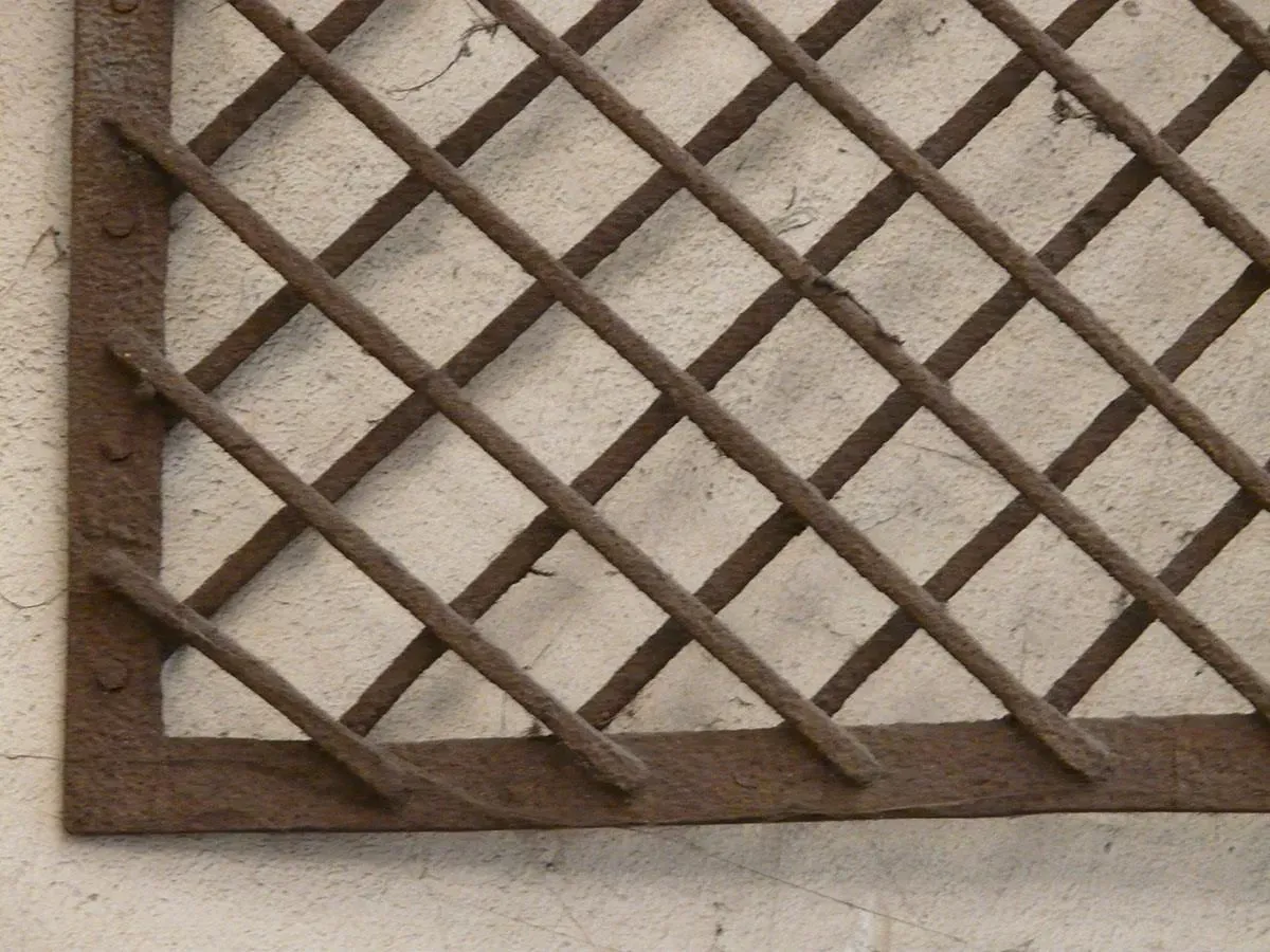 Rostiges Metallgitter in Rautenmuster vor einer cremefarbenen Wand.