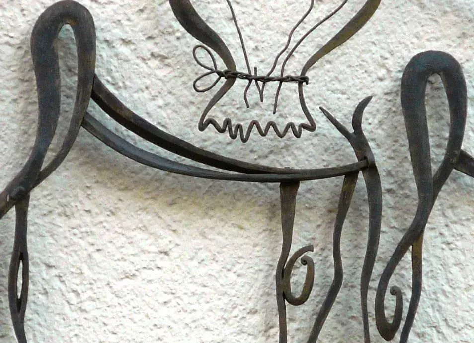 Metallskulptur einer Figur mit Rüschenkragen und gewundenen Gliedmaßen vor einer Stuckwand.