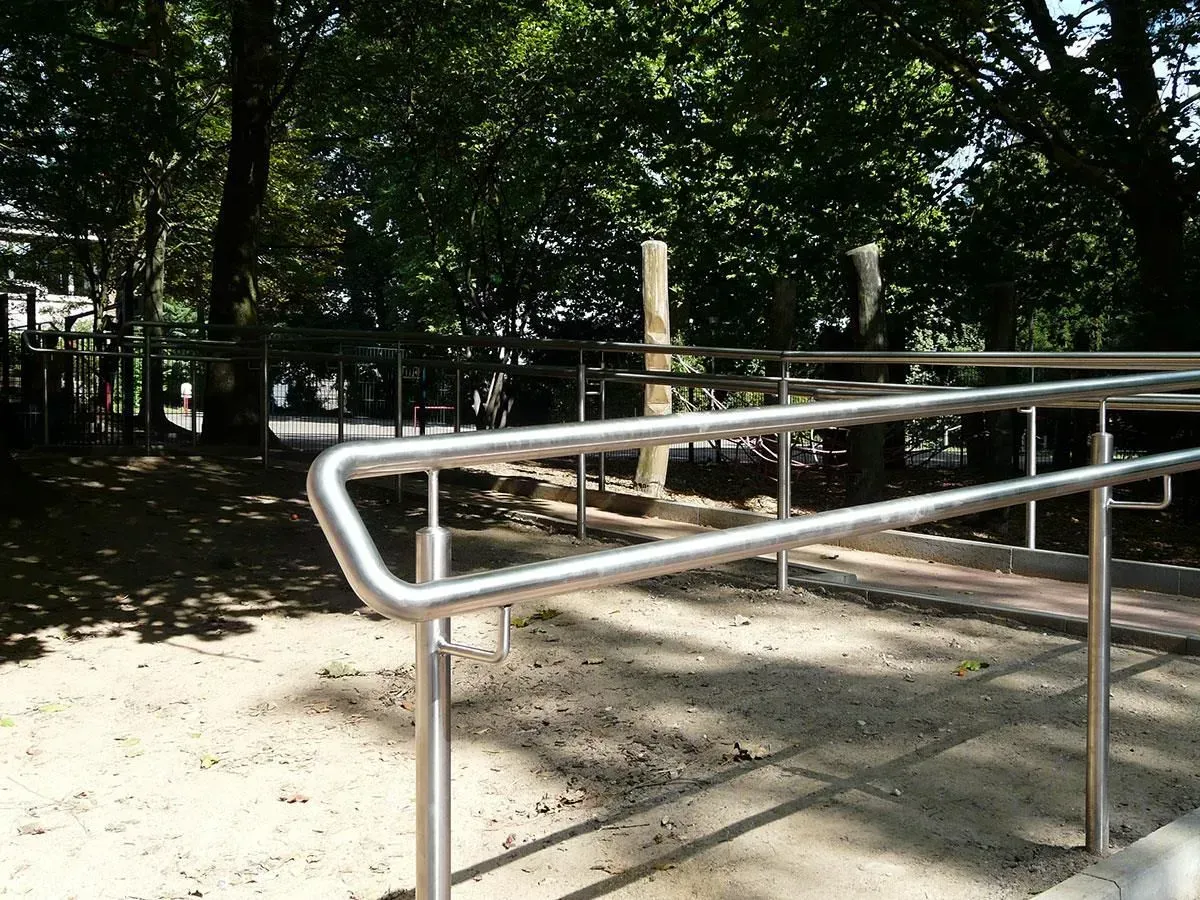 Handläufe an einer Rampe, die in eine Parkanlage mit Bäumen und einem Steinweg führt.