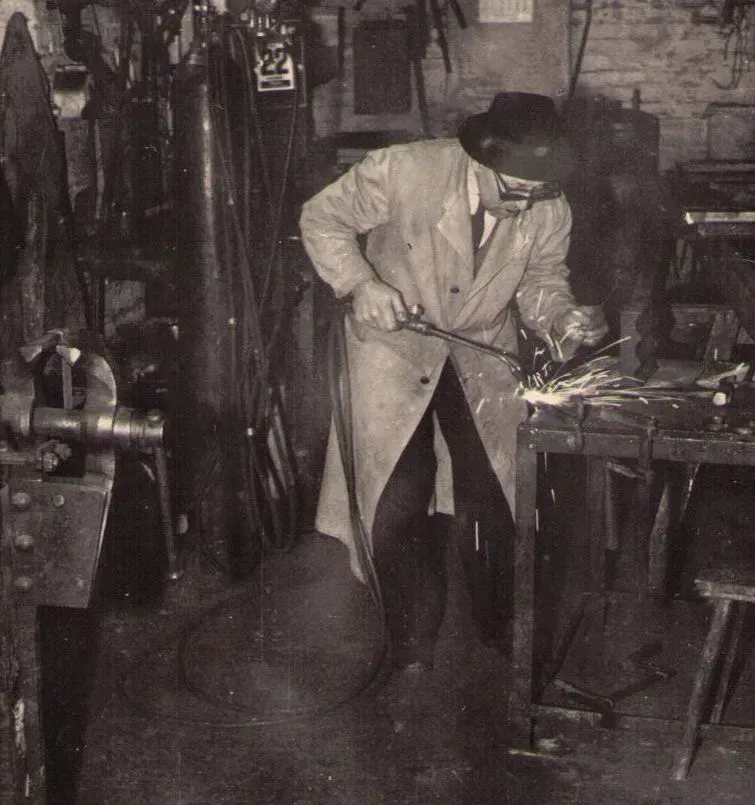 Mann mit Hut und Mantel schweißt Metall in einer überfüllten Werkstatt, Funken fliegen.