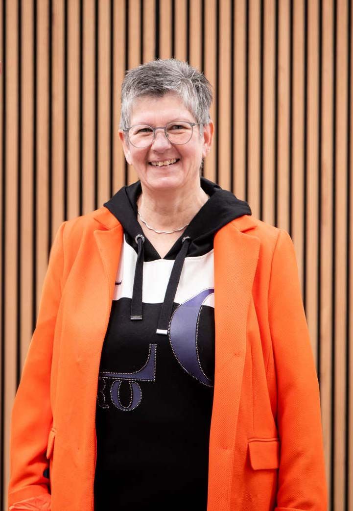 Eine Frau in einer orangefarbenen Jacke und einem schwarzen Hemd lächelt vor einer Holzwand.
