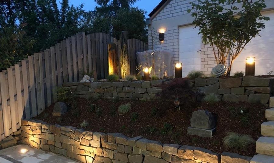 Vor einem Haus ist nachts eine Steinmauer beleuchtet.