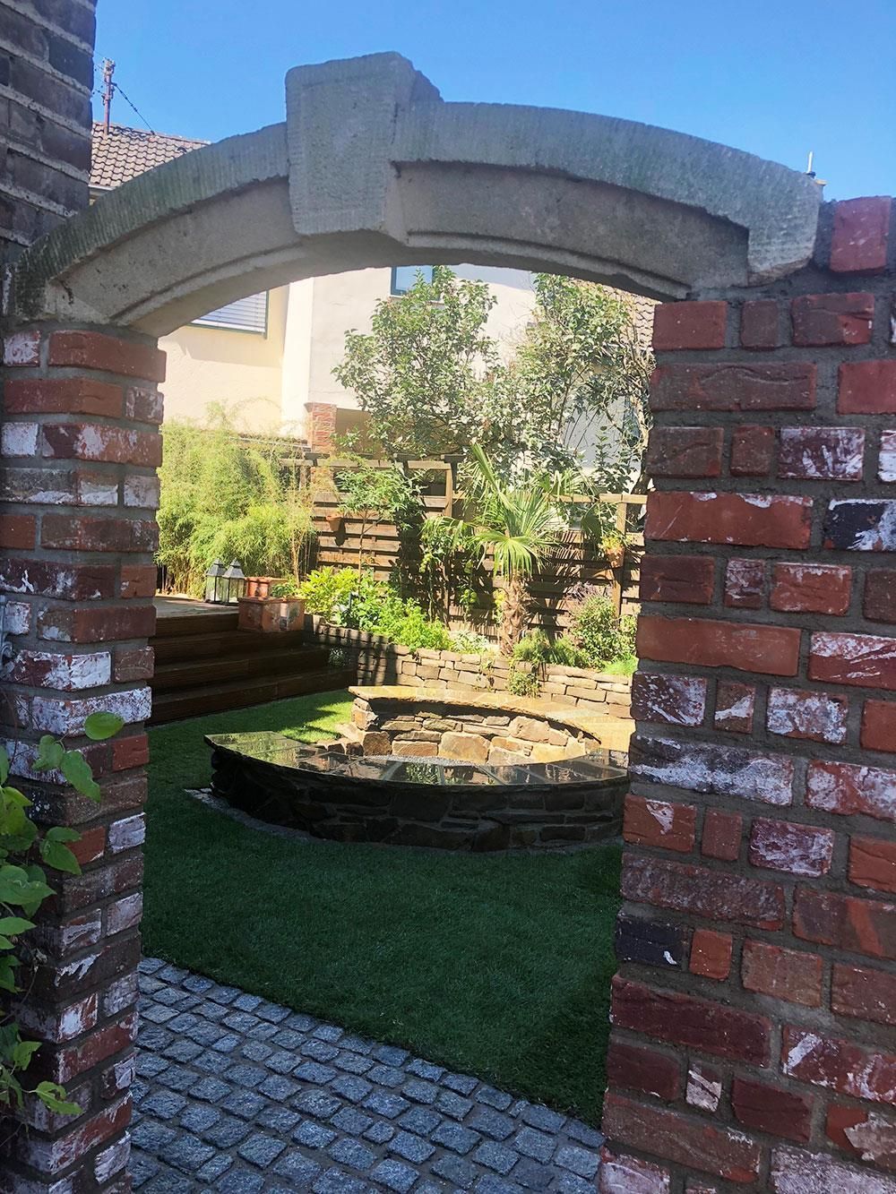 Eine Backsteinmauer mit einem steinernen Torbogen, der zu einem Garten führt.