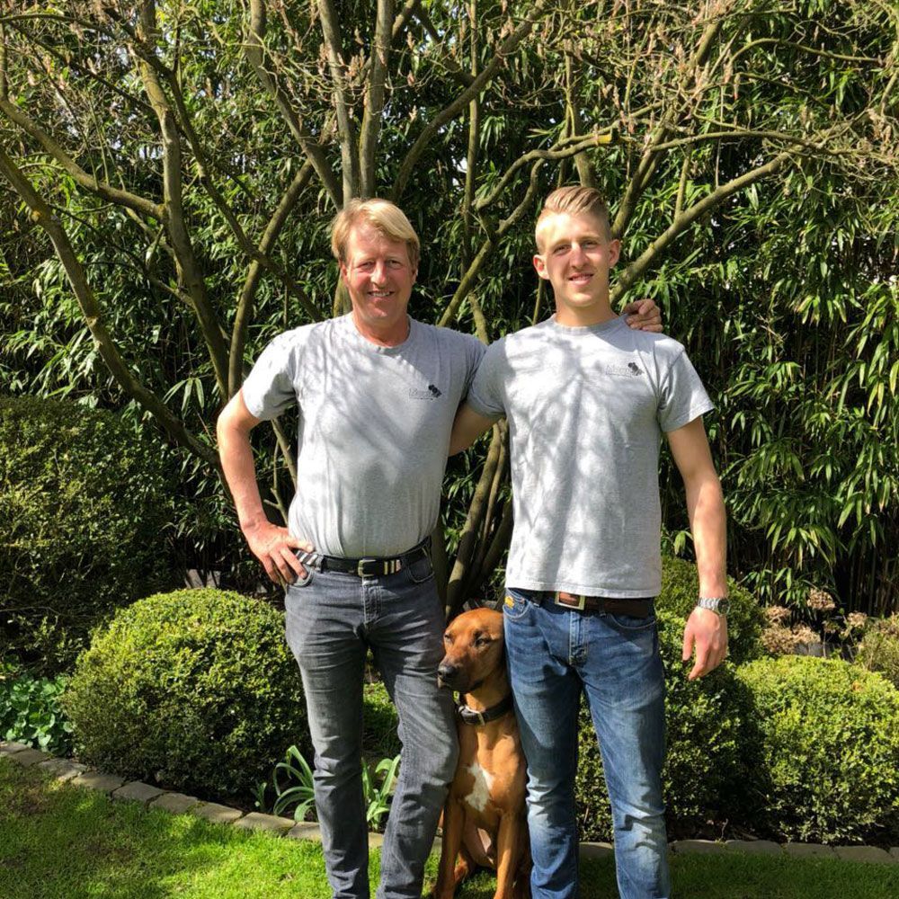 Zwei Männer stehen neben einem Hund in einem Garten.