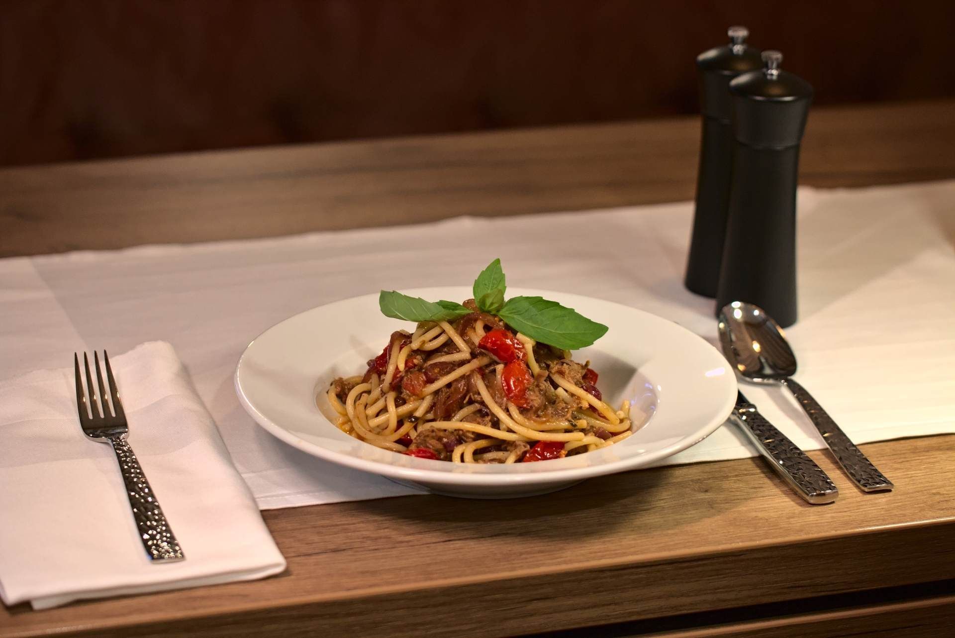 Spaghetti mit Tomatensosse, garniert mit Basilikum, serviert auf einem weissen Teller mit Besteck, Serviette und Pfeffermühlen.