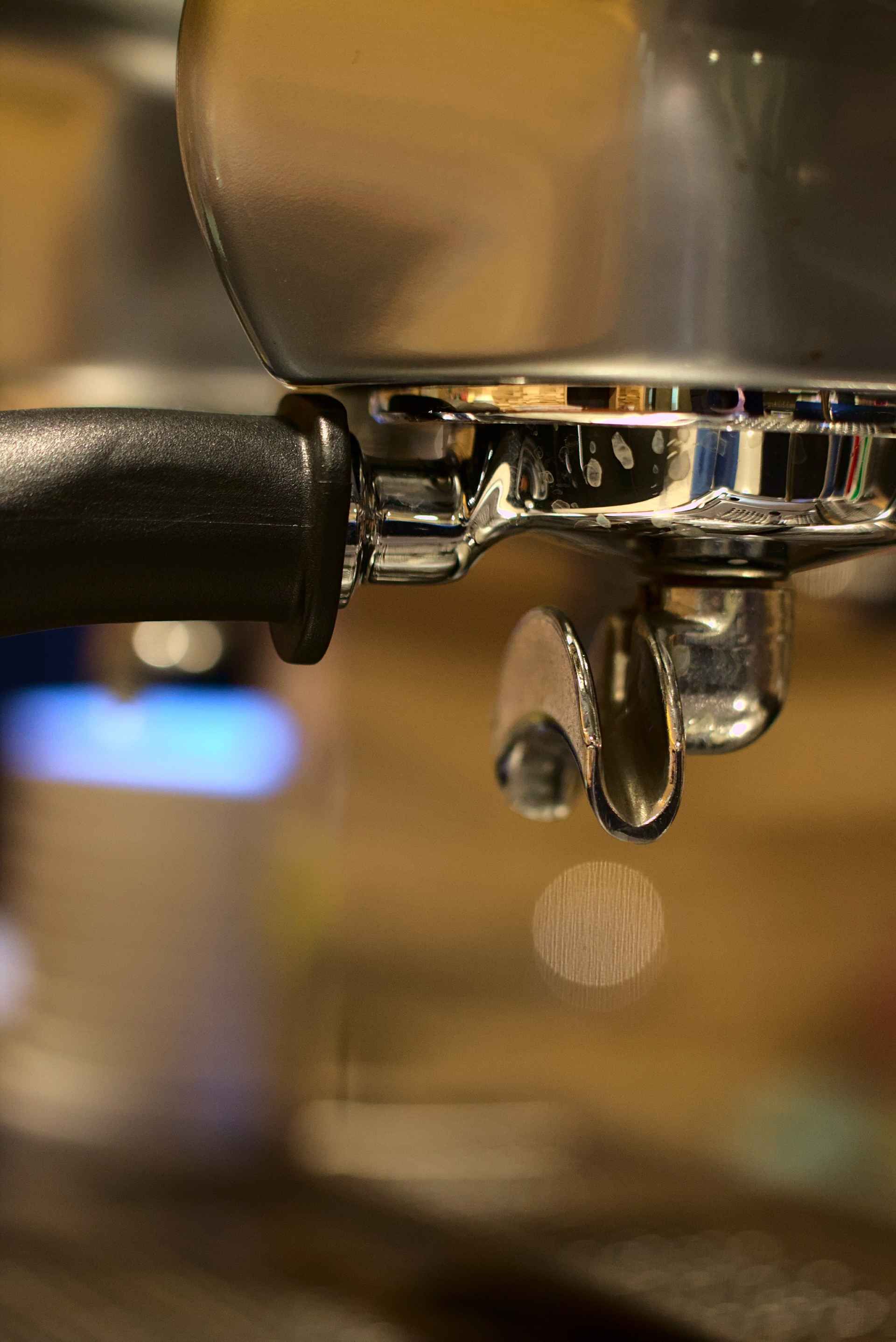 Nahaufnahme eines verchromten Siebträgers einer Espressomaschine mit einem einzelnen Tropfen Flüssigkeit. Schwarzer Griff, unscharfer Hintergrund.