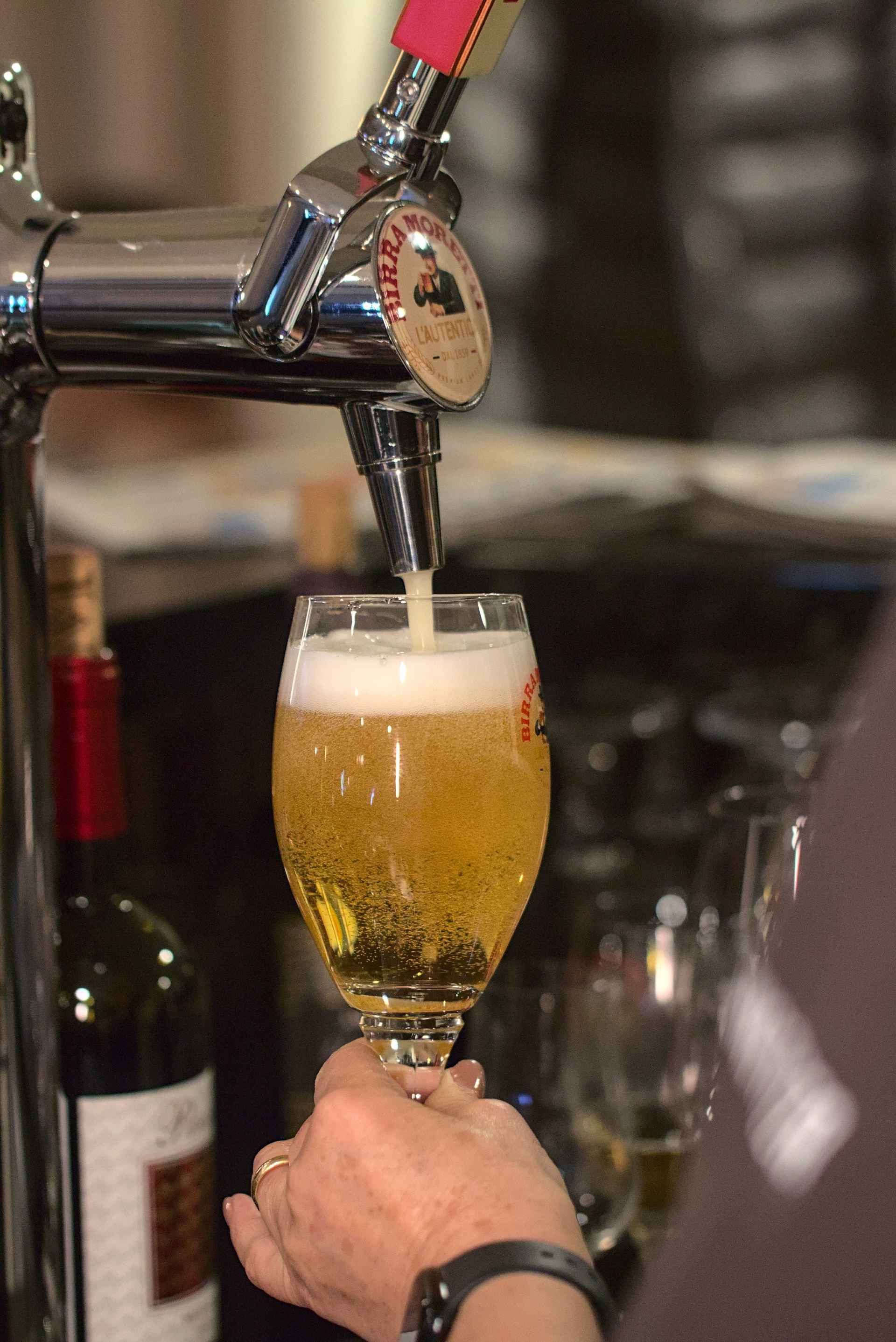 Bier wird vom Zapfhahn in ein Glas gezapft, das von einer Hand gehalten wird. Im Hintergrund steht eine Weinflasche.