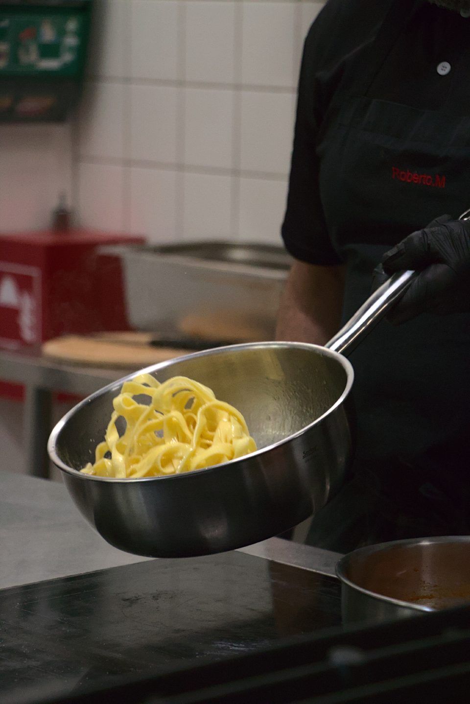 Ein Koch hält in einer Küche einen Topf mit gekochten Fettuccine-Nudeln in der Hand.