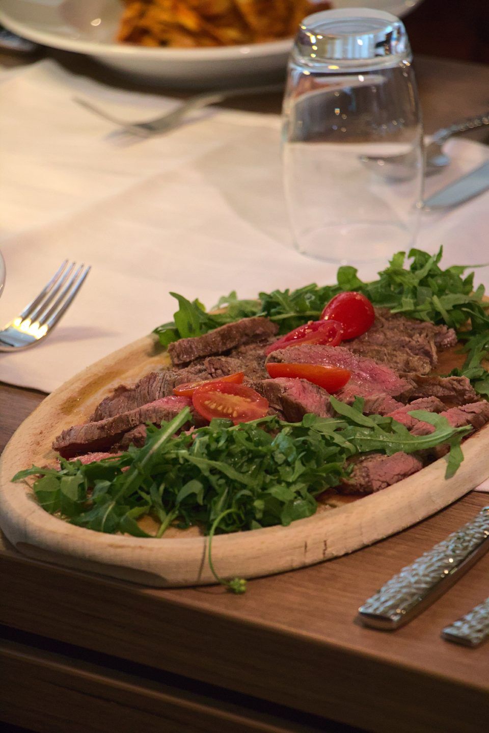 Geschnittenes Steak auf einem Holzbrett mit Rucola und Tomaten, serviert an einem Restauranttisch.