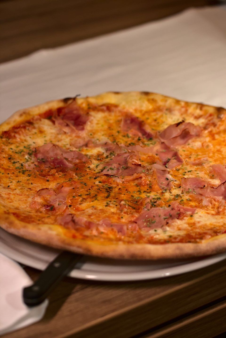 Pizza mit Schinken und Kräutern auf einem weissen Teller, Holztisch.