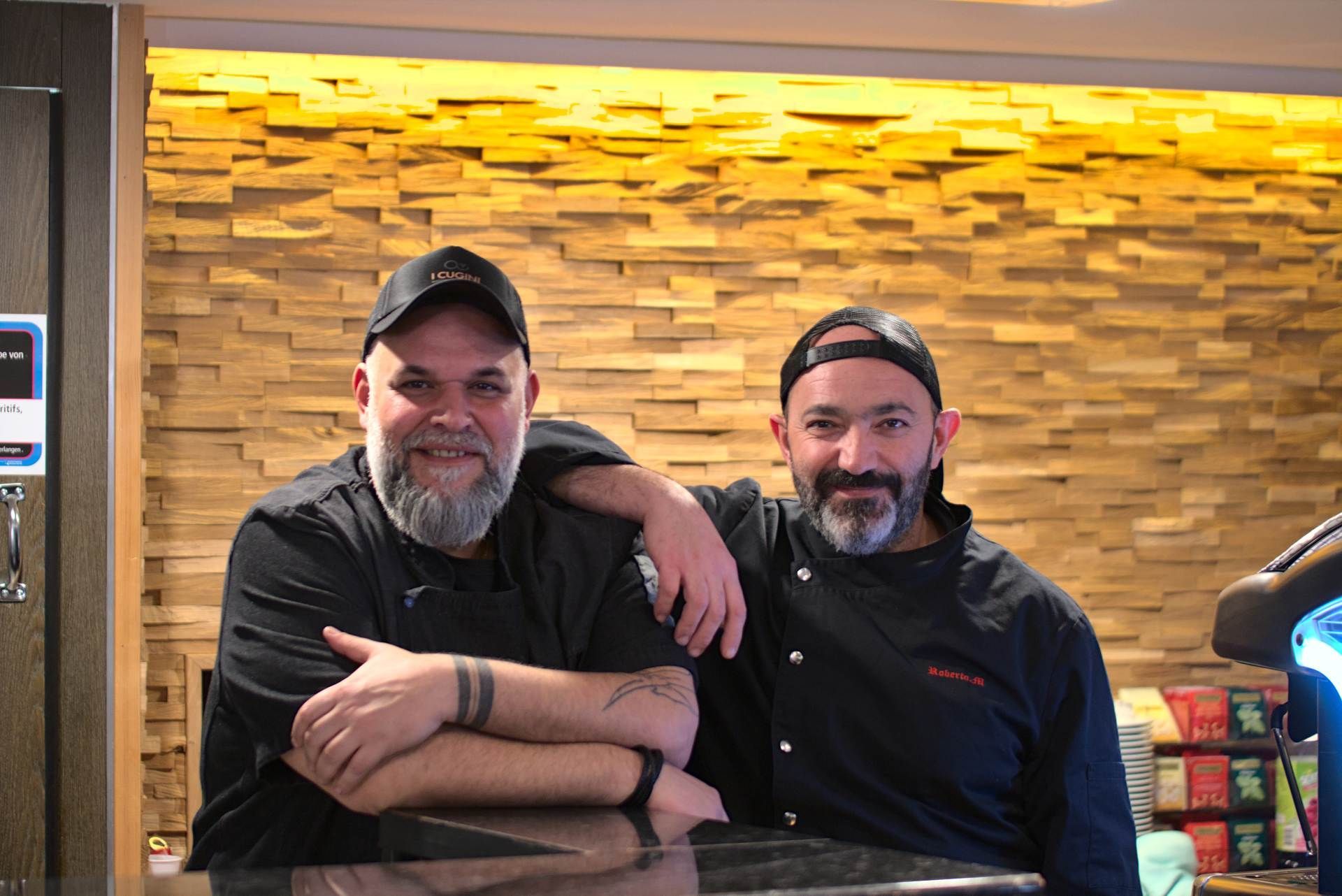 Zwei Männer in schwarzen Kochjacken und -mützen lächeln, einer hat den Arm um die Schulter des anderen gelegt, vor einer holzgetäfelten Wand.