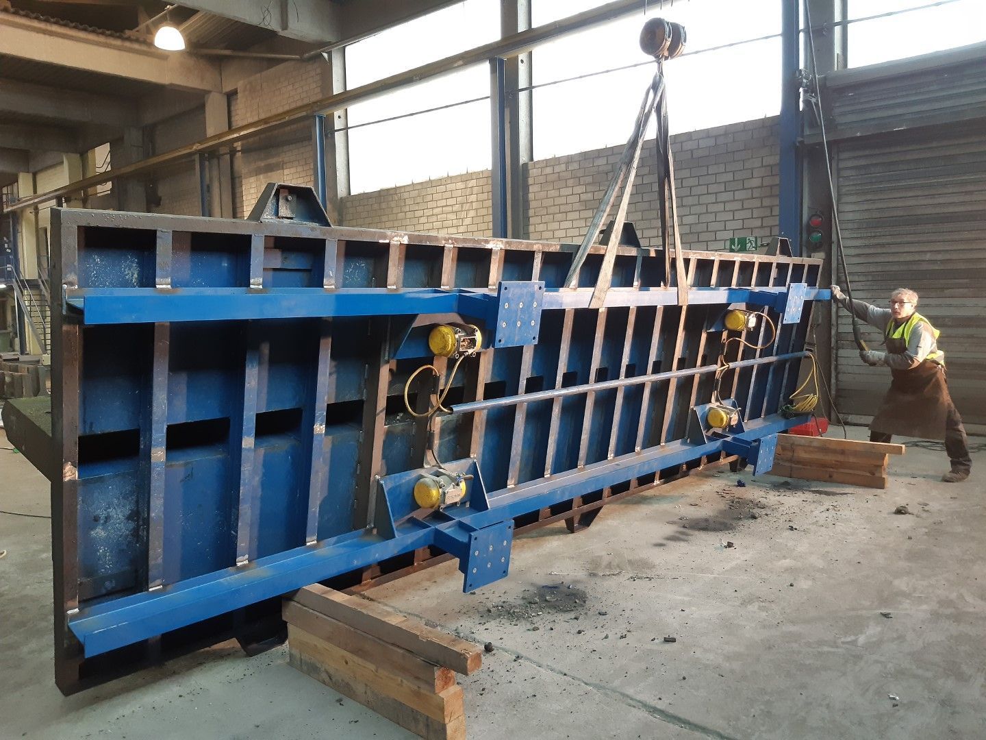 Eine große blaue Metallkonstruktion wird in einer Werkstatt von einem Kran angehoben; ein Arbeiter steht in der Nähe.