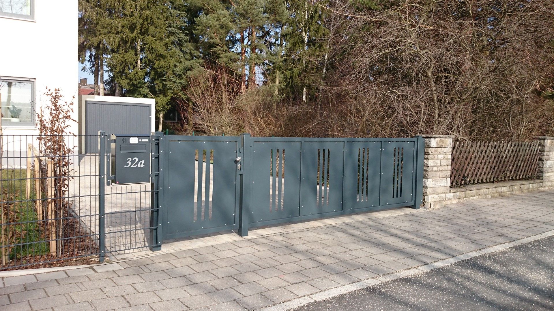 Graues Metalltor mit dekorativen Ausschnitten führt zu einem Haus auf einem Ziegelweg.